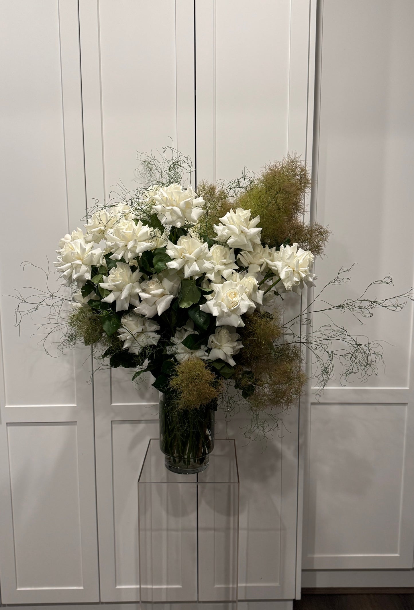 vase of large white blooming roses surrounded by various greenery against a white backdrop on a glass pedestal