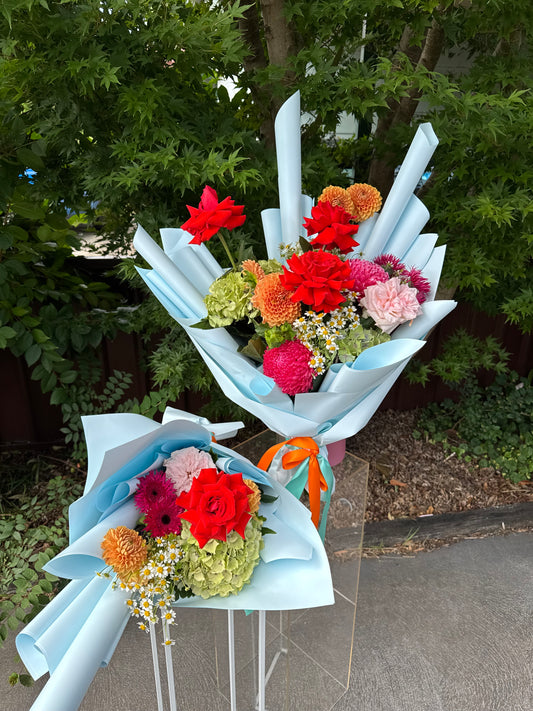 A personalized floral arrangement with a mix of red and green flowers, tied with blue paper and ribbons, displayed outdoors.